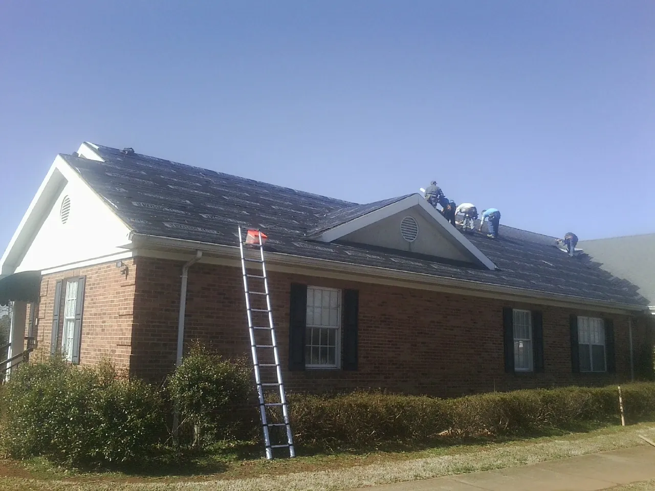 Crew tearing shingles off a brick ranch roof with a ladder set — Metal Roof Repair work by our crew in Waubun