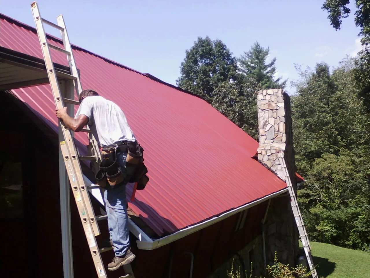 Roofer on a ladder beside a red metal roof and stone chimney — Soffit & Fascia Repair by Roofing Brothers in Waubun